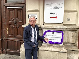 Behindertenanwalt Dr. Hansjörg Hofer vor dem Eingang mit einem violett verkleideten Schild der Behindertenanwaltschaft